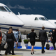 A group of six people walking in front of a row of private jets on the tarmac.